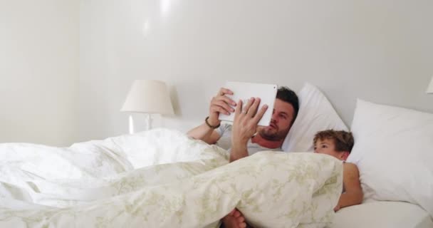 Lad os tage et hurtigt kig, før vi står op. 4k video optagelser af en ung smuk far og hans søn ved hjælp af en tablet i sengen derhjemme. – Stock-video