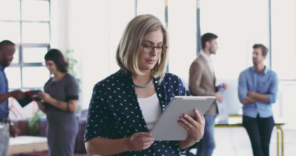 Siempre sobresalen de la multitud. Imágenes de video 4K de una mujer joven usando una tableta digital mientras está de pie en una oficina moderna con compañeros de trabajo en el fondo. — Vídeos de Stock