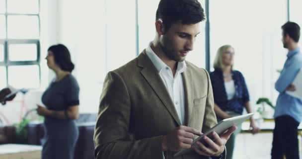 Haciendo negocios. Imágenes de video 4K de un joven usando una tableta digital mientras está de pie en una oficina moderna con compañeros de trabajo en el fondo. — Vídeo de stock