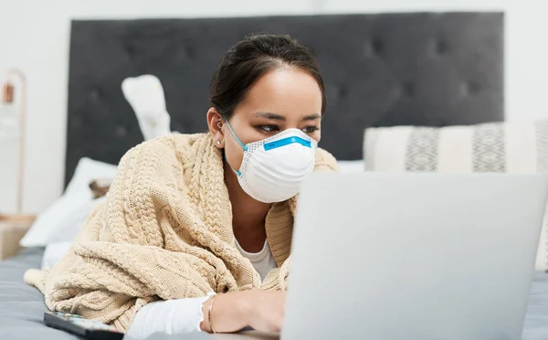 Immer auf dem Laufenden mit den neuesten Gesundheitsratschlägen. Aufnahme einer Frau mit Maske, während sie mit ihrem Laptop auf ihrem Bett liegt. — Stockfoto