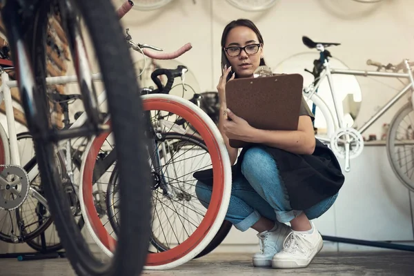 Biały czy różowy? Całkowite ujęcie atrakcyjnej młodej kobiety kucającej w swoim sklepie rowerowym i korzystającej z telefonu komórkowego trzymając schowek. — Zdjęcie stockowe