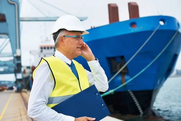 ¿Dónde está mi cliente? Un trabajador portuario de pie en el puerto en medio de la actividad de la industria naviera mientras recibe una llamada en su móvil. — Foto de Stock