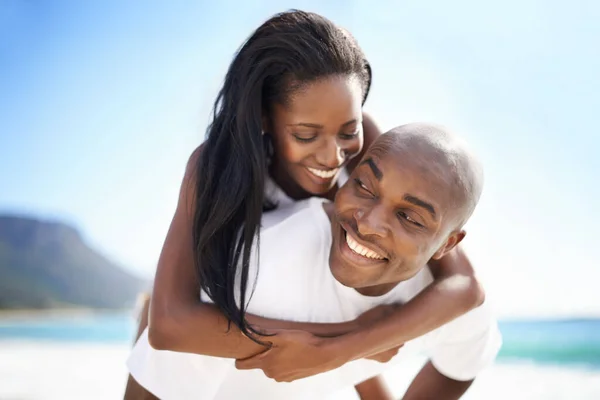 Surpresa, meu amor. Um homem afro-americano bonito dando a sua namorada um piggyback na praia. — Fotografia de Stock