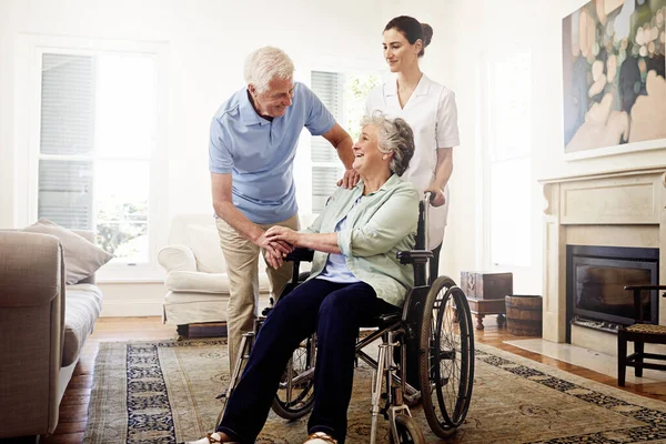 A vida de um finalista. Tiro de um cuidador sorridente com uma mulher idosa em uma cadeira de rodas e seu marido em casa. — Fotografia de Stock