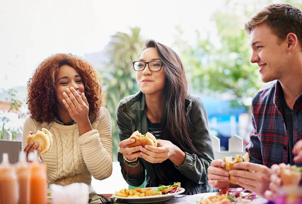 Dobré jídlo a smích jdou ruku v ruce. Vystřižený záběr tří přátel jíst hamburgery venku. — Stock fotografie