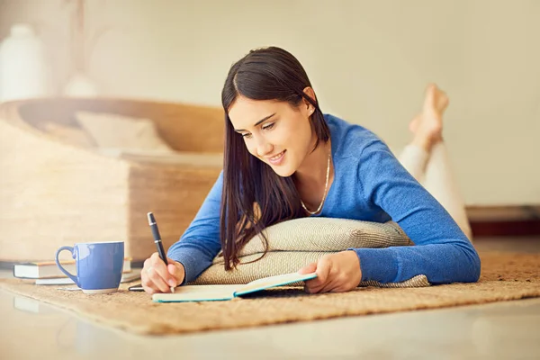 Jag skriver bara ner några tankar. Skjuten av en glad ung kvinna gör anteckningar i sin anteckningsbok när hon ligger på sin lounge golv. — Stockfoto