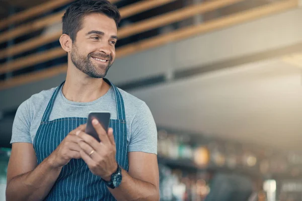 Los baristas inteligentes usan la tecnología más inteligente. Fotografía de un joven usando un teléfono móvil mientras trabajaba en una cafetería. — Foto de Stock