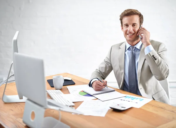 Manejando tus activos como si fuera el suyo. Retrato de un joven empresario hablando por teléfono mientras escribía en su escritorio. — Foto de Stock