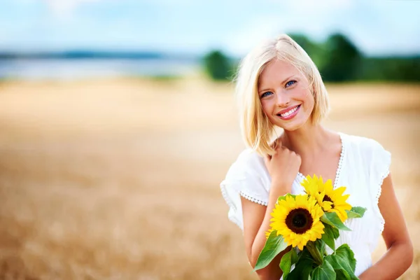 Ela é uma beleza natural em seu elemento. Linda jovem mulher sorrindo em um campo de trigo, enquanto segurando alguns girassóis. — Fotografia de Stock