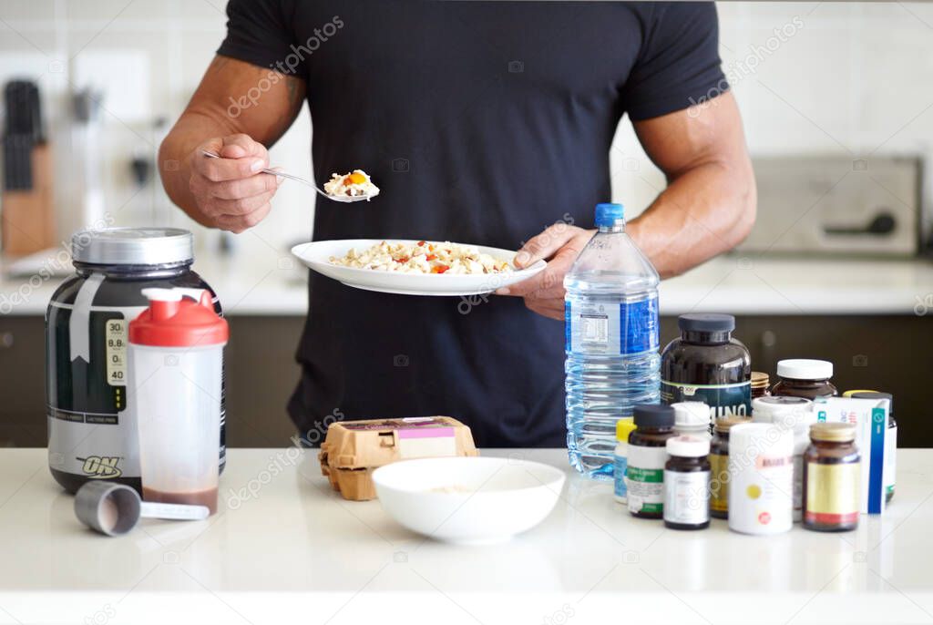 Making sure he has all his vitamins and minerals. A muscular man holding a bowl of omelet while standing in front of his supplements.