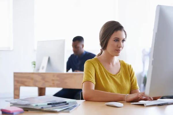 Eficiência é tudo. Uma jovem mulher trabalhando em sua mesa com seu colega em segundo plano. — Fotografia de Stock