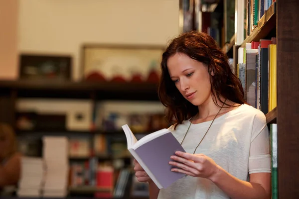 Absorbed in the plot. A young woman lost in the pages of a novel against a backdrop of full bookshelves. — Stock Photo, Image