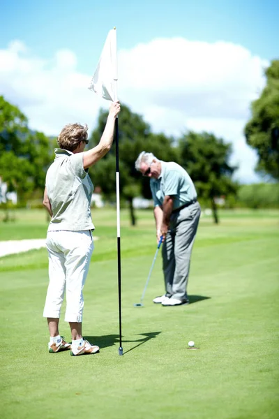 Master of the hole-in-one. A senior woman holding a flag while her husband puts for birdie. — Stock Photo, Image