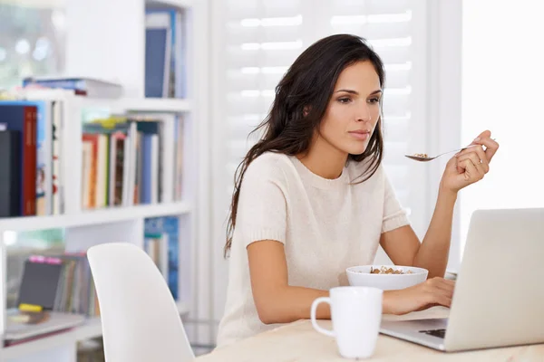 Online all the time. Shot of a busy young woman eating breakfast while surfing the net. — Stock Photo, Image