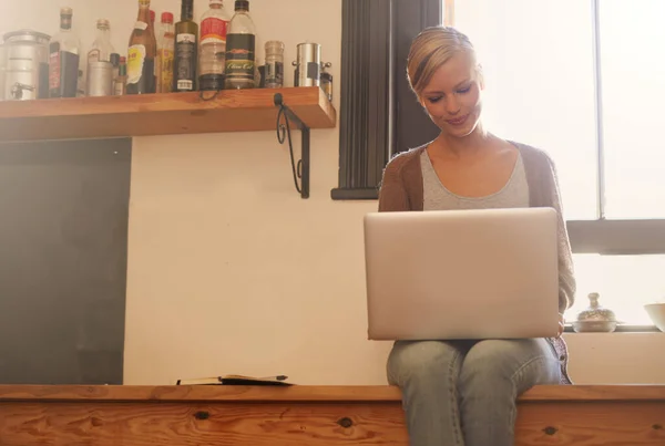 Die Küche ist ihr Lieblingsplatz, um alles zu erledigen. Eine attraktive junge Frau benutzt ihren Laptop, während sie auf ihrem Küchentisch sitzt. — Stockfoto
