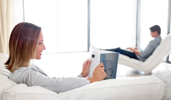 Even bijpraten over de laatste mode. Een jonge vrouw leest een tijdschrift terwijl haar vriend bladert een laptop op de achtergrond. — Stockfoto