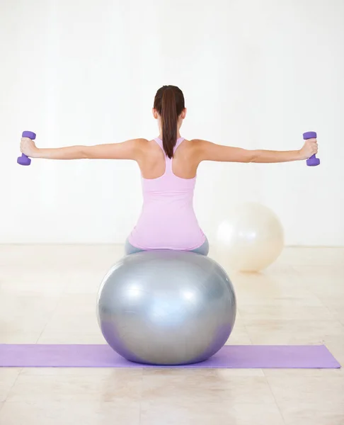 Два основных компонента ее тренировки. Пересмотр снимка молодой женщины, сидящей на мяче для упражнений и выполняющей упражнения с гантелями. — стоковое фото