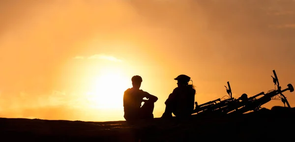 Her şeyin güzelliğini göz önünde bulundurarak gün batımını izleyen bir çiftin siluetini sahilde yanlarında bisikletleriyle izliyorlar.. — Stok fotoğraf