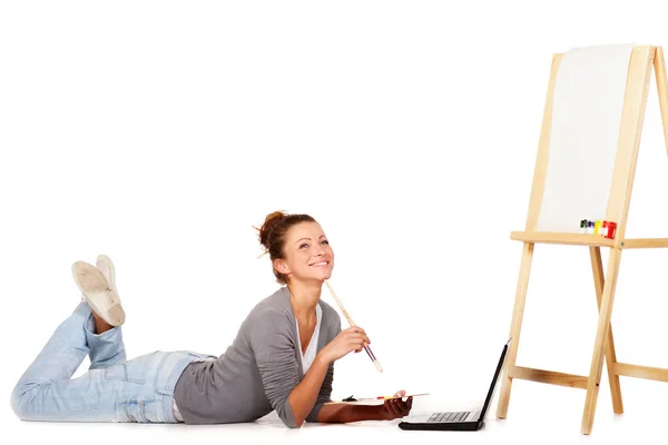Wszędzie szukam inspiracji. Uśmiechnięta młoda kobieta leżąca przy laptopie i puste płótno na sztaludze. — Zdjęcie stockowe