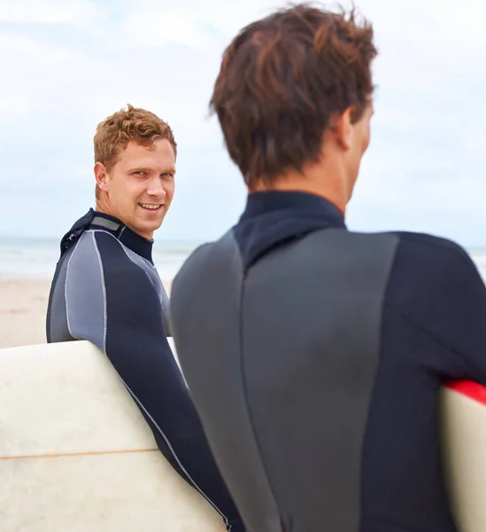 Vinculados por su amor al surf. Dos jóvenes surfistas de pie en la playa con el océano al fondo. — Foto de Stock