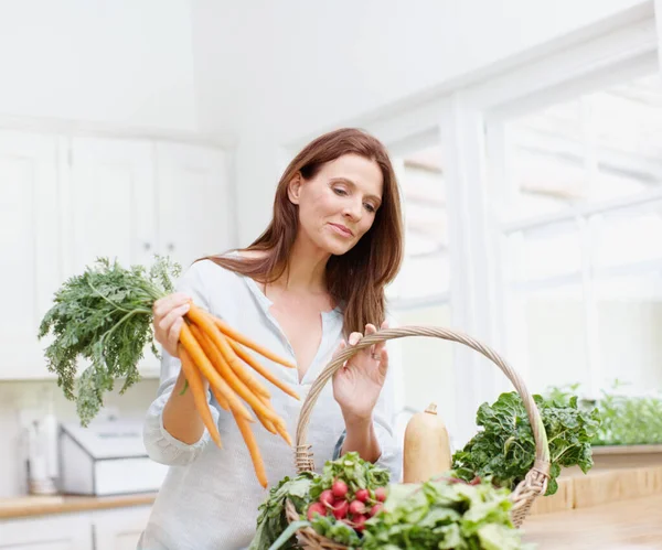 Sopivat ainekset hienoon minestronikeittoon. Upea nainen pitää kasan porkkanoita katsoessaan hänen koriinsa vihanneksia keittiössään.. — kuvapankkivalokuva
