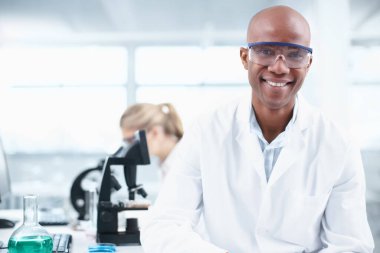 Gülümseyen araştırmacı. Arka planda bayan bir iş arkadaşıyla laboratuvarda gülümseyen genç bir kimyagerin portresi..