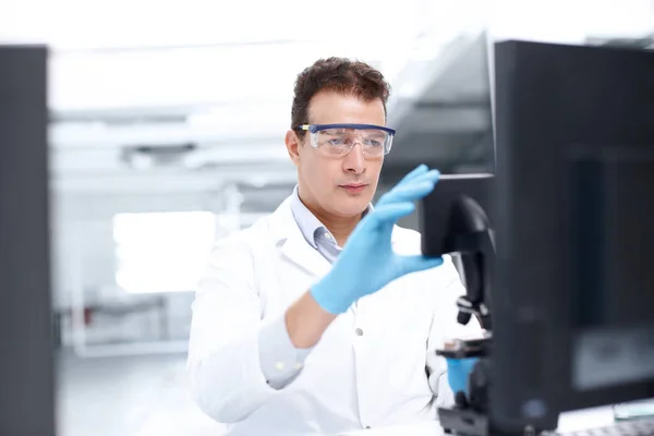 Göra justeringar. En vetenskapsman tittar uppmärksamt på ett mikroskop medan han bär skyddsglasögon. — Stockfoto