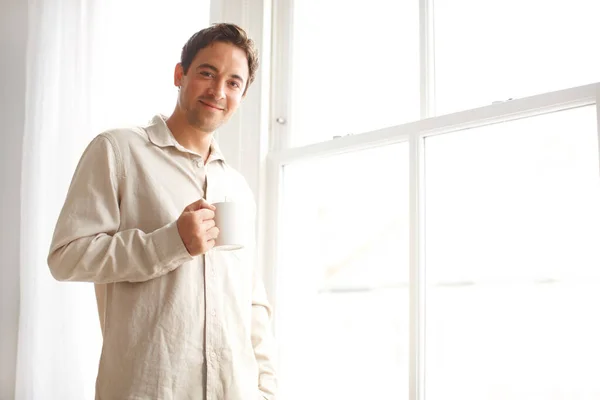 Aspettiamo una buona giornata. Hansome giovane uomo in piedi vicino alla finestra della sua camera da letto mentre tiene una tazza di caffè. — Foto Stock