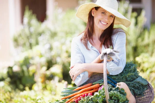 A kertészkedés életem szerelme. Egy gyönyörű nő szalmakalapban, kezében egy kosár zöldséggel a kertjében.. — Stock Fotó