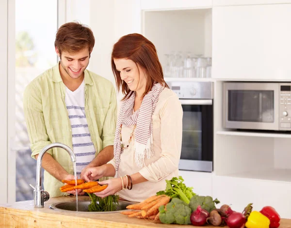 Posso lavar estas cenouras, se quiseres. Um casal feliz lavando algumas cenouras na pia da cozinha. — Fotografia de Stock