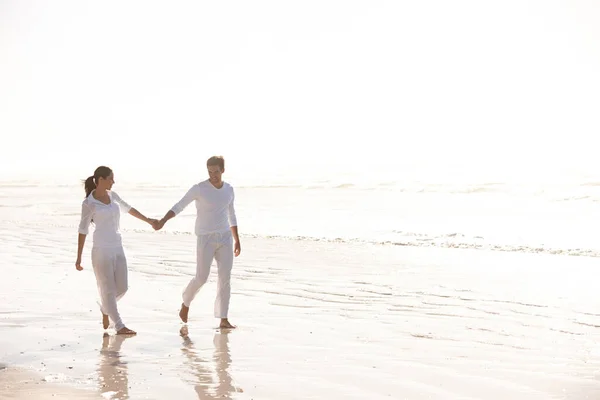 Se tenir la main à travers la beauté de la nature. Plan complet d'un beau jeune couple vêtu de blanc marchant le long d'une plage. — Photo