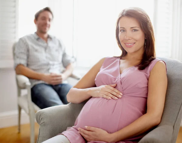 Getting ready for parenthood. Portrait of a beautiful pregnant woman holding her stomach while her husband sits in the background. — Stock Photo, Image