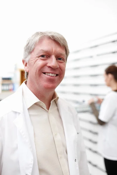 Mosolygós, érett gyógyszerész. Egy jóképű, érett gyógyszerész portréja, aki a háttérben dolgozó kollégájával mosolyog. — Stock Fotó