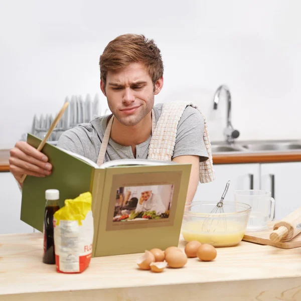 Dit is een beetje moeilijker dan ik had verwacht.... Een jongeman die in zijn keuken staat en verbaasd kijkt terwijl hij een receptenboek leest. — Stockfoto