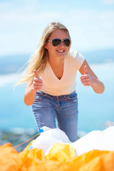 Jump success. A beautiful young woman giving two thumbs up outdoors with a parachute in the foreground. — Stock Photo, Image