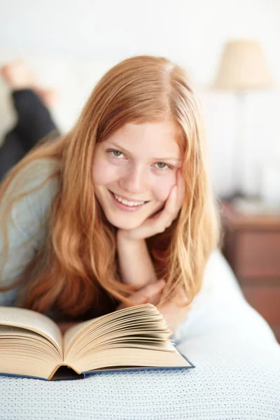 Наслаждаюсь миром вымысла. Портрет молодой рыжей женщины, читающей книгу и лежащей на кровати. — стоковое фото
