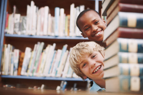 Hello. Two school friends peering around a stack of books in the library.