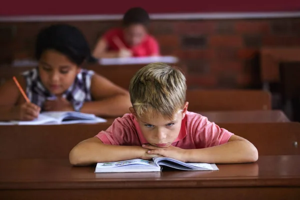 School is klote. Een jonge jongen rust zijn hoofd op zijn armen als hij zit in een klaslokaal kijkt verveeld. — Stockfoto