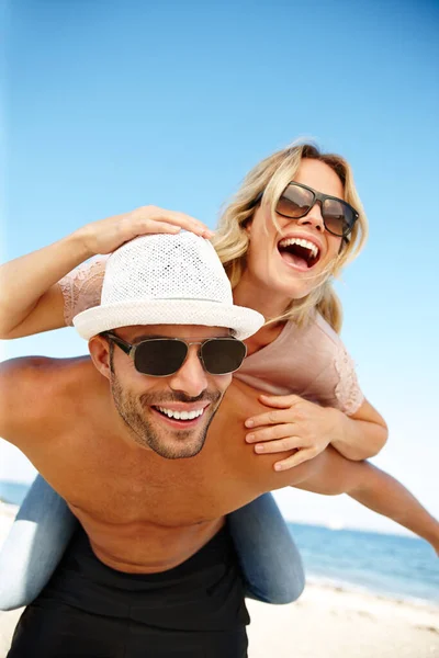 Leuke vakantiedagen. Schot van een lachende jongeman die zijn lachende vriendin een meelift geeft op een zonnig strand. — Stockfoto