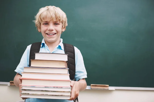 Ik wil mijn hersenen zo vol mogelijk inpakken. Een leuke jonge jongen met een stapel boeken tegen een schoolbord in de klas. — Stockfoto