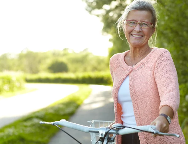 Lubi zdrowe zajęcia na świeżym powietrzu - jazda na rowerze. Uśmiechnięta starsza kobieta stojąca na wiejskim pasie rowerowym. — Zdjęcie stockowe