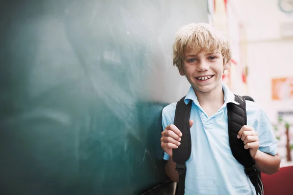 他准备好学了。一个快乐的小男孩站在黑板旁的版权空间旁边，手里拿着背包的带子. — 图库照片