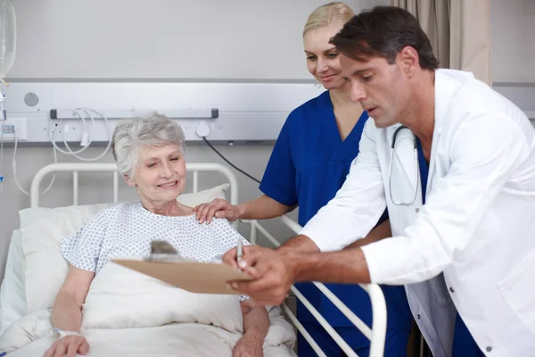 Son buenas noticias. Un médico y su joven enfermera le muestran dónde firmar el informe médico mientras está de pie sobre su cama. —  Fotos de Stock