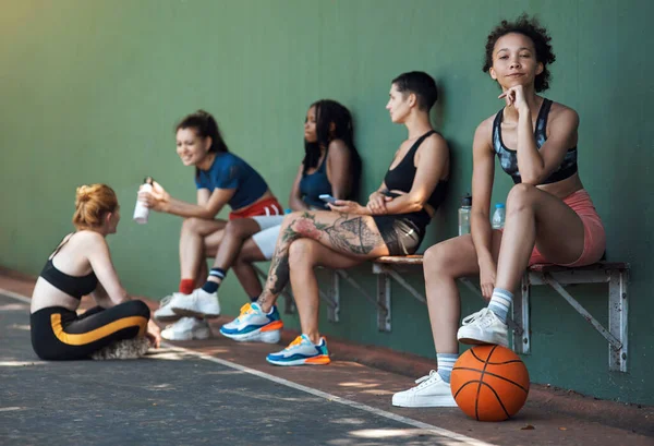 Creo que tienes lo que se necesita. Retrato completo de una atractiva joven atleta sentada en un banco en la cancha de baloncesto con sus compañeras de equipo en el fondo. — Foto de Stock
