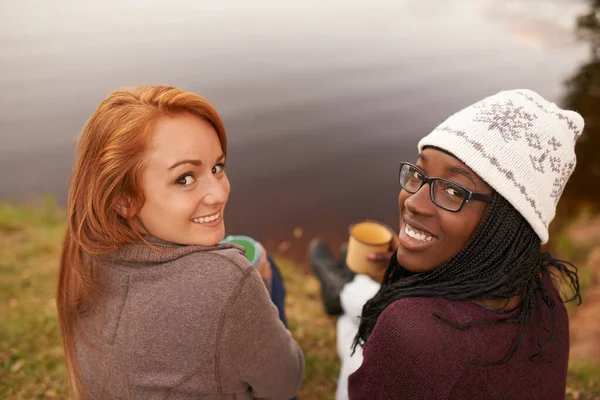 Passeios de inverno para o lago. Retrato de alto ângulo de duas adolescentes felizes sentadas ao lado de um lago. — Fotografia de Stock