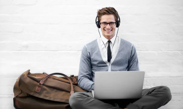 Esperar pelo teu voo não tem de ser chato. Um jovem homem de negócios bonito sentado no chão usando fones de ouvido enquanto digita em seu laptop com bagagem ao lado dele. — Fotografia de Stock