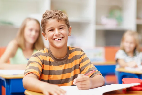 Наслаждаюсь беззаботным моментом в классе. Милый школьник, сидящий за столом в классе и счастливо смеющийся.. — стоковое фото