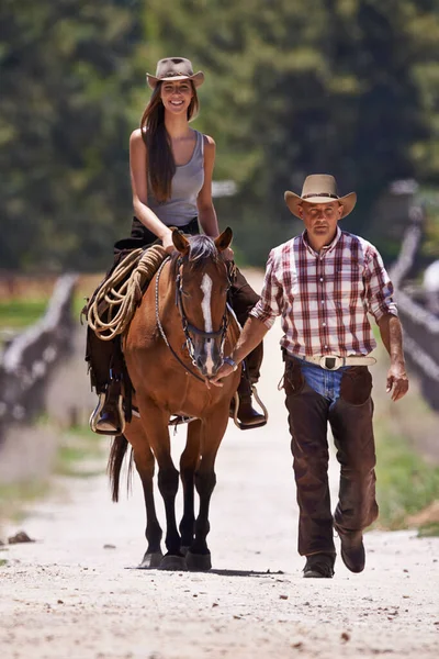 Cabalgando 101. Una atractiva joven vaquera montando un caballo en un rancho mientras un vaquero camina junto a. —  Fotos de Stock