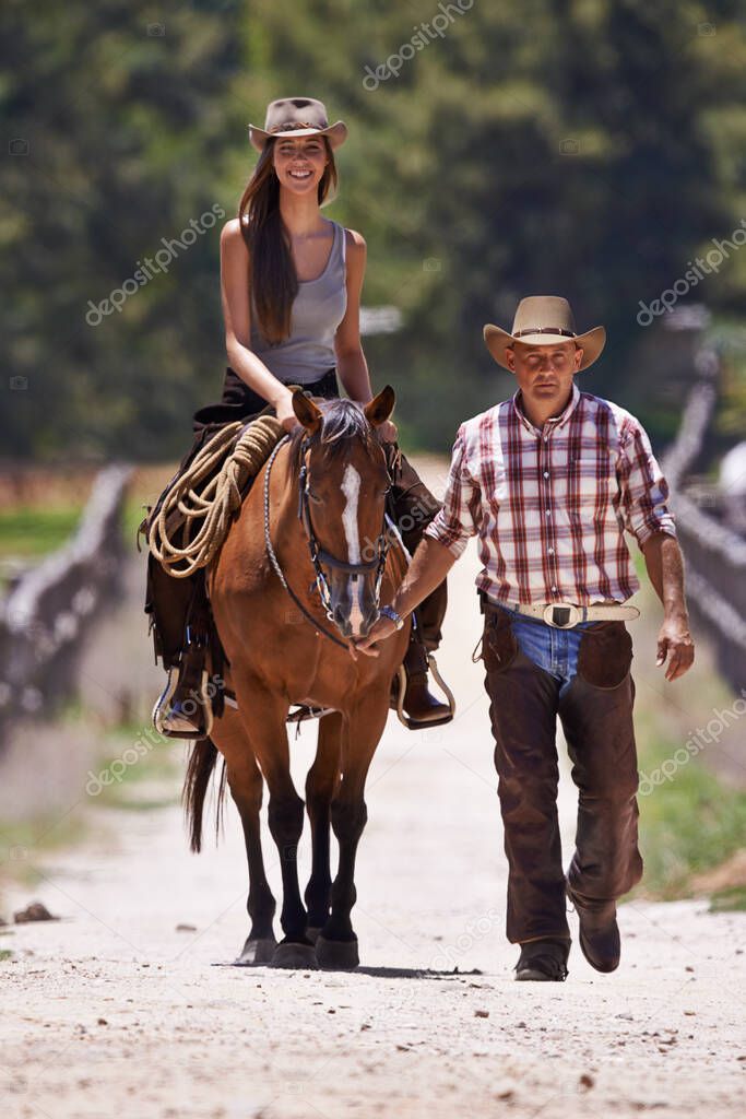 Riding 101. An attractive young cowgirl riding a horse on a ranch while a cowboy walks alongside.