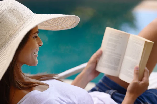 The perfect holiday read. Shot of a young woman reading a book by the poolside. Stock Picture